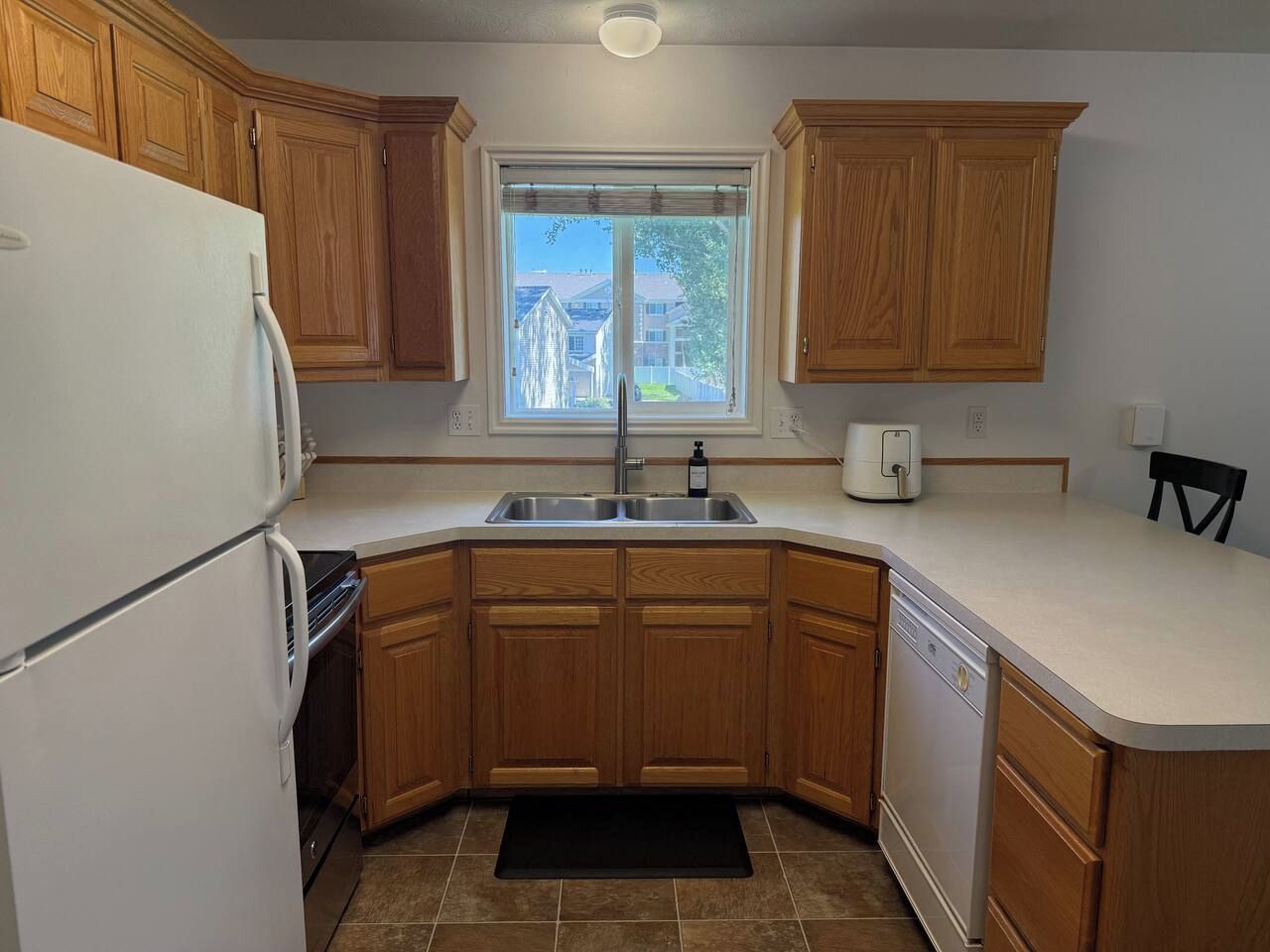 A kitchen with wooden cabinets, a white refrigerator, a sink, and a window with a view of houses.