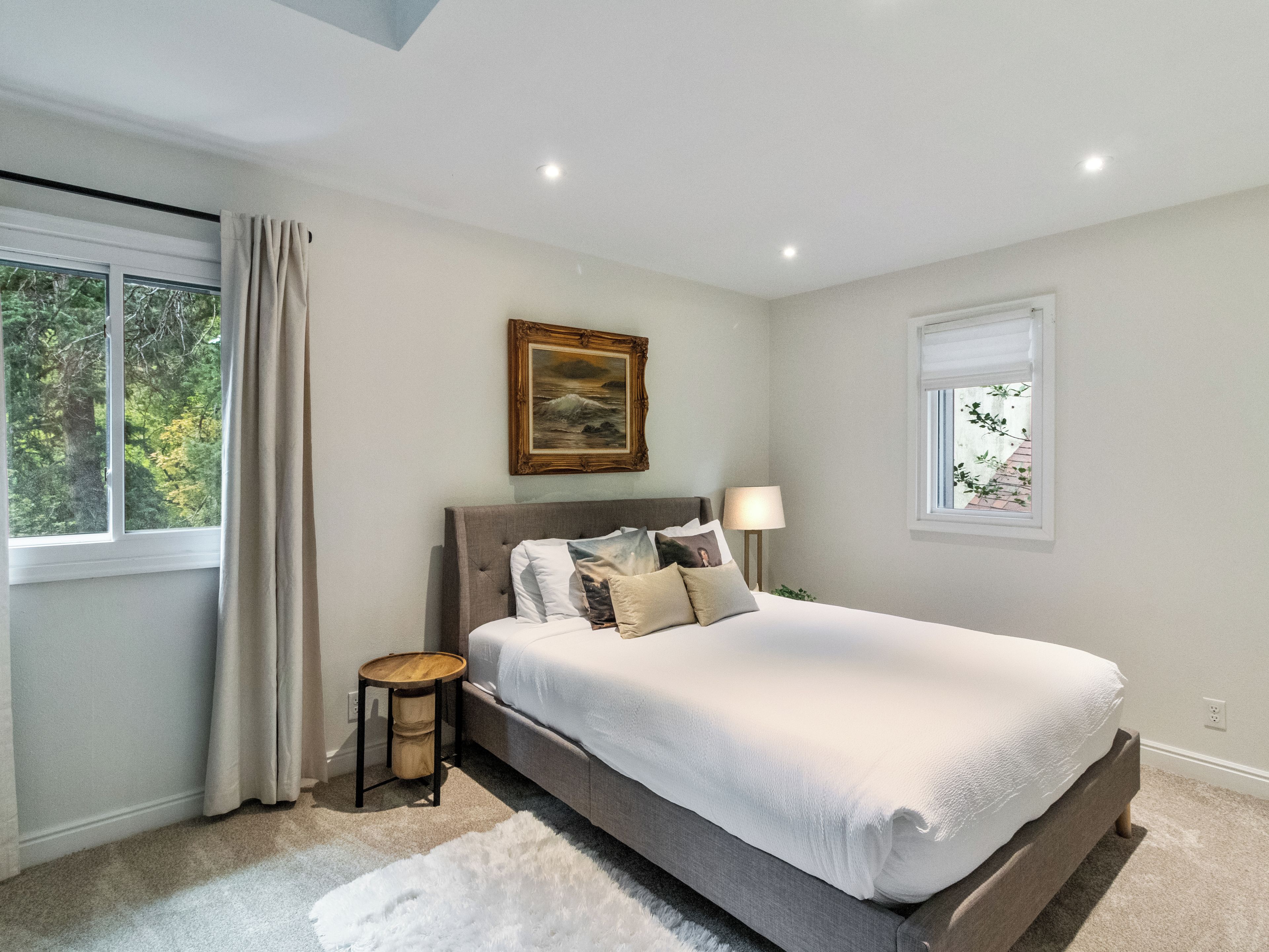 Un dormitorio con una cama grande, una mesita de noche, una lámpara, un cuadro y una ventana con cortinas.