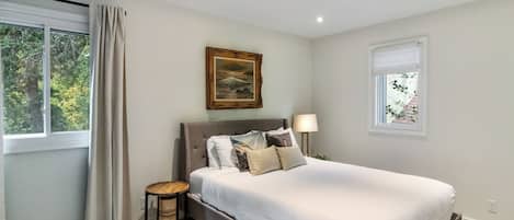 Un dormitorio con una cama grande, una mesita de noche, una lámpara, un cuadro y una ventana con cortinas.