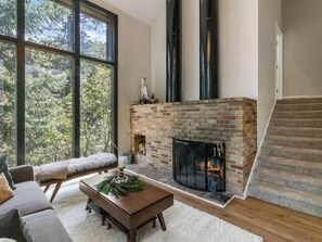 A spacious living room with a brick fireplace, large windows, a wooden staircase, and a cozy seating area.