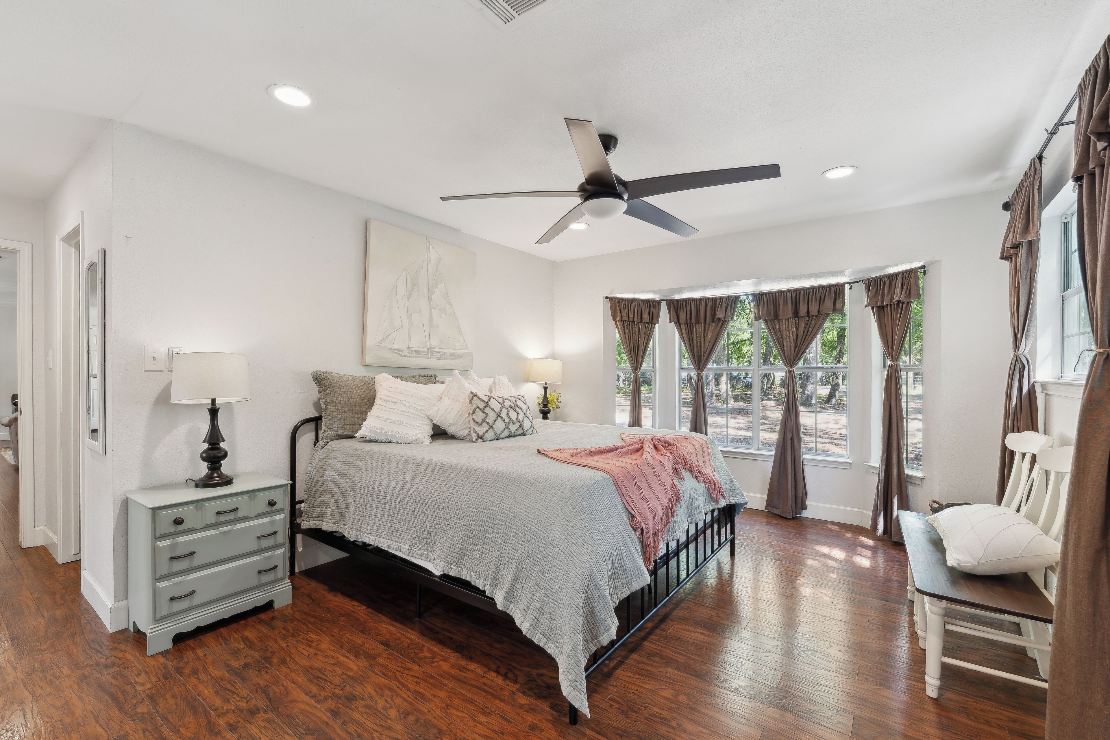 A bedroom with a bed, bedside table, lamp, ceiling fan, and large windows with curtains.