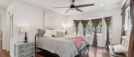 A bedroom with a bed, bedside table, lamp, ceiling fan, and large windows with curtains.