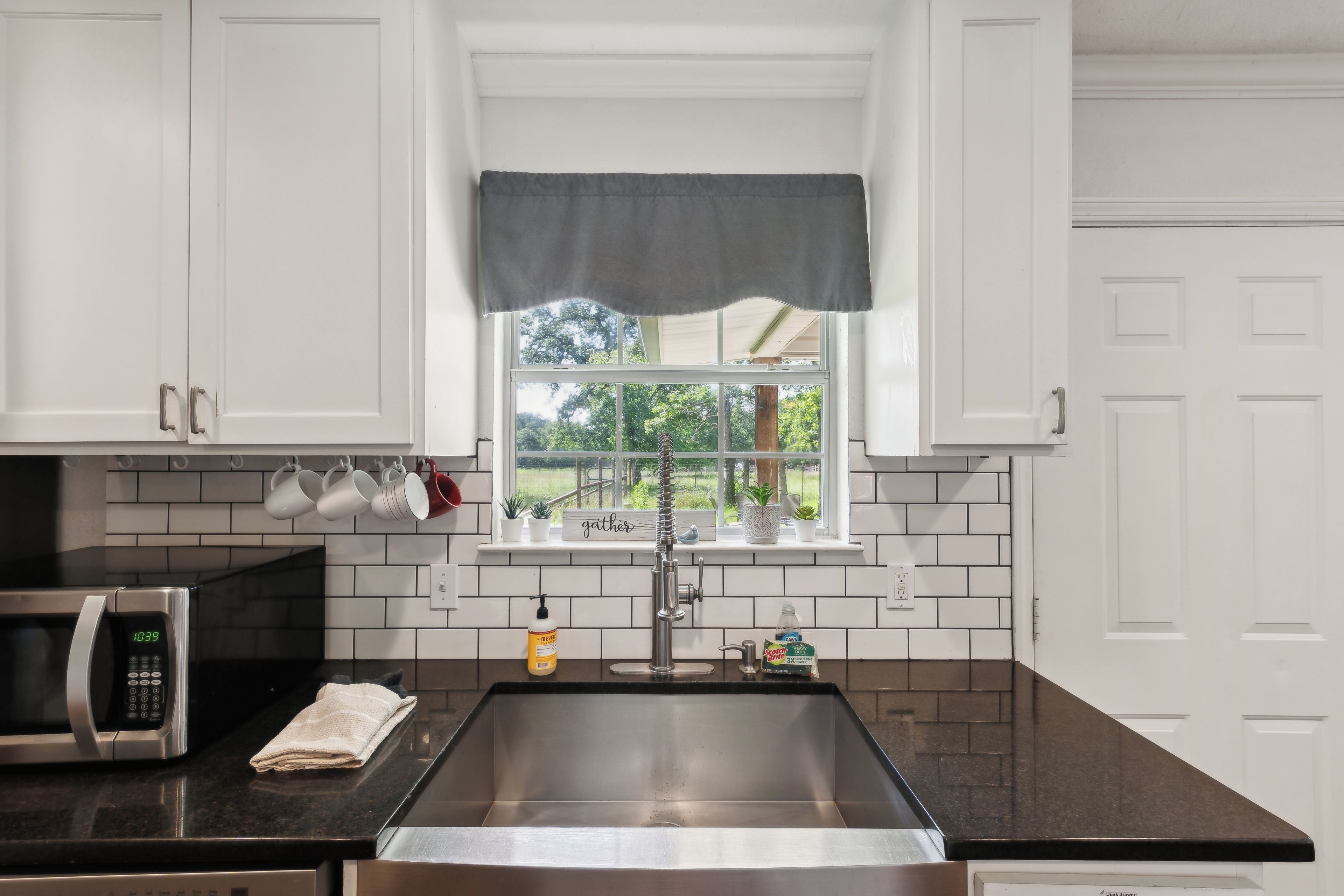 A kitchen with white cabinets, a stainless steel sink, a microwave, and a window with a view of greenery.