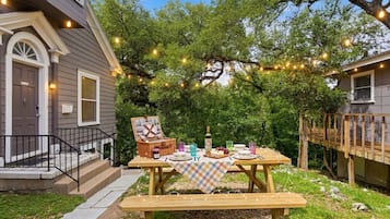 A picnic setup with a table, benches, and string lights in a backyard with trees and a house.
