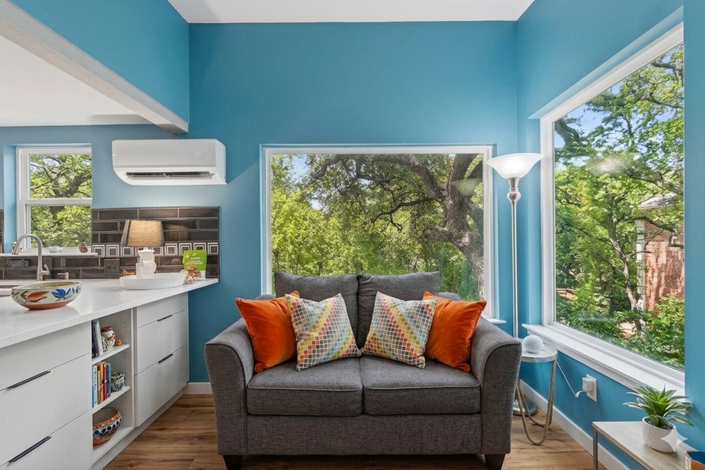 Ein Zimmer mit grauem Sofa, einem Fenster mit Blick auf Bäume, einer weißen K üchenzeile und einer blauen Wand.