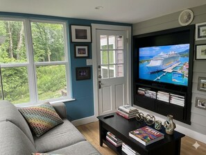 Ein Wohnzimmer mit großem Fenster, Fernseher, einer Couch, einem Couchtisch mit Büchern und Deko sowie einer Tür.