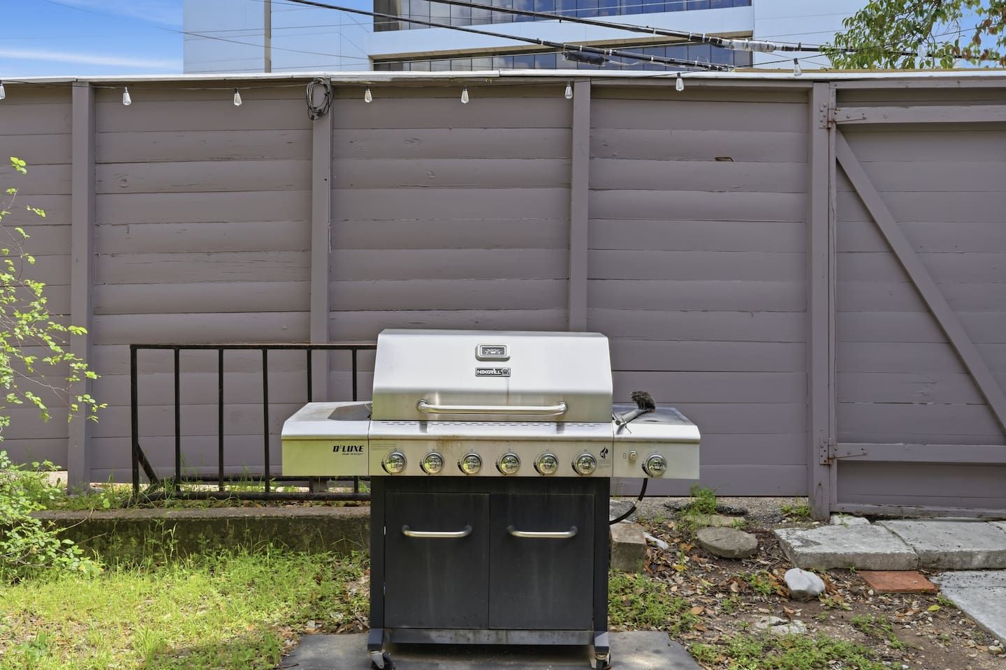 A D'Luxe outdoor grill with a control panel, a storage cabinet, and a metal railing in the background.
