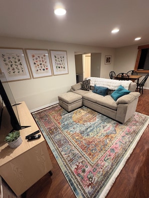 Un salon avec un canapé d’angle, un tapis à motifs, une table basse agrémentée d’une plante et de télécommandes, et des reproductions botaniques encadrées accrochées au mur.