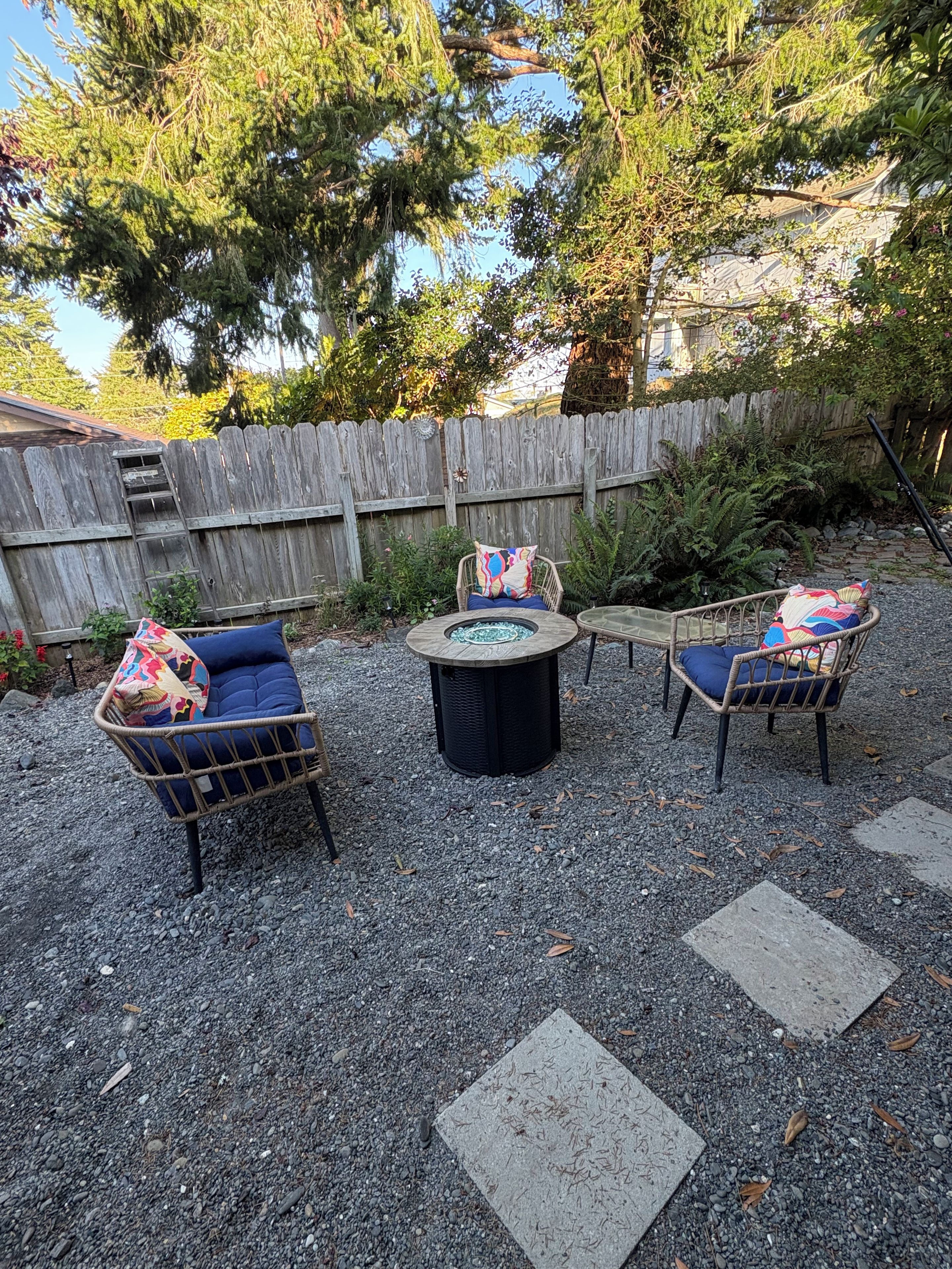 An outdoor seating area with wicker chairs and a round table, surrounded by gravel and a wooden fence.