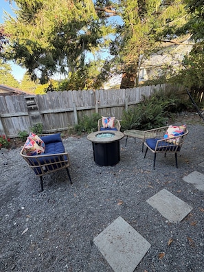 Un espace extérieur aménagé avec des chaises en osier et une table ronde, entouré de gravier et d’une clôture en bois.