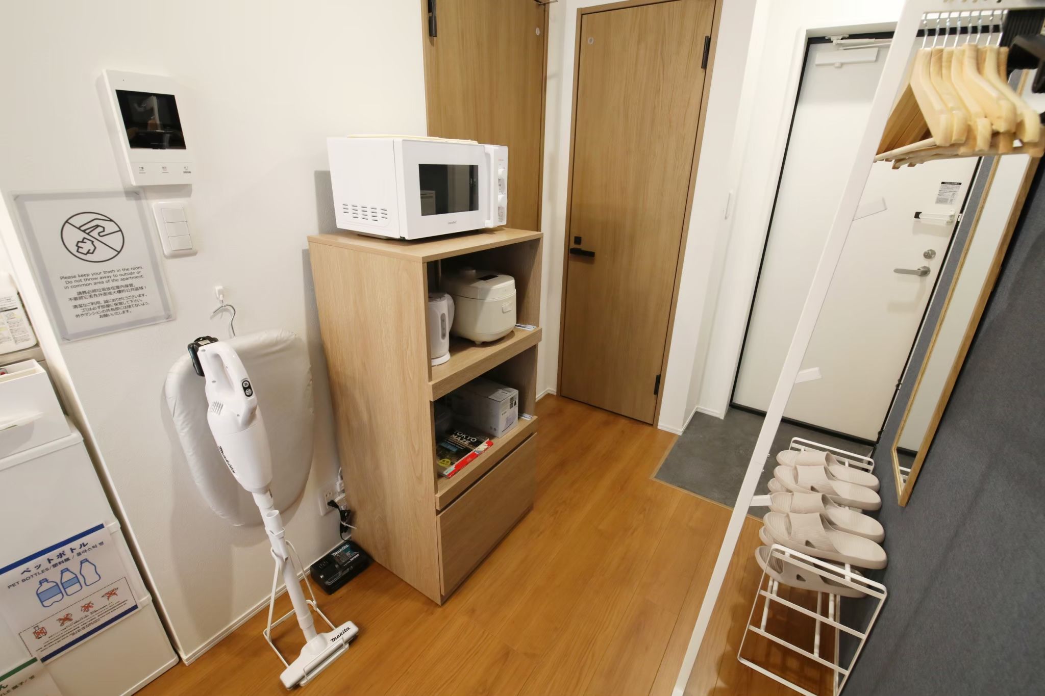 A compact hotel room with a microwave, a small refrigerator, a vacuum cleaner, a wooden cabinet with storage, and a shoe rack.