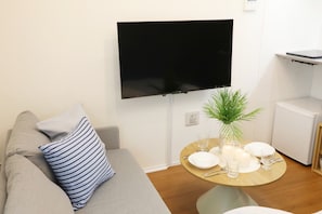 A living room with a flat-screen TV mounted on the wall, a round wooden dining table set with plates, glasses, and a candle, and a grey sofa with striped pillows. - Lamarie Haneda 402 (Ota Ku)