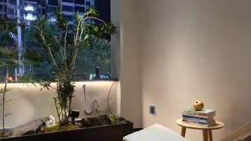 Um quarto com cama, uma mesinha com livros, uma janela com vista para prédios e um arranjo de plantas.