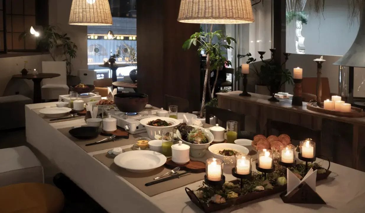 Uma sala de jantar bem iluminada com uma mesa comprida posta para uma refeição, velas e vista para o exterior.