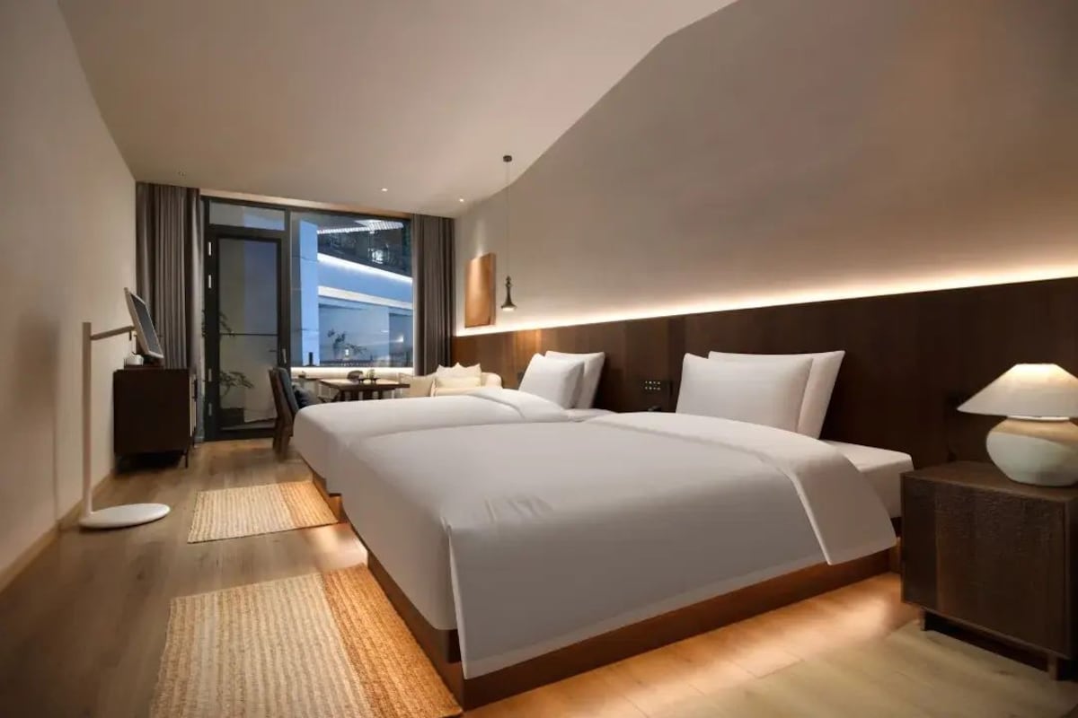 Quarto de hotel moderno com uma cama grande, mesas de cabeceira, uma escrivaninha e vista através de uma porta de vidro.