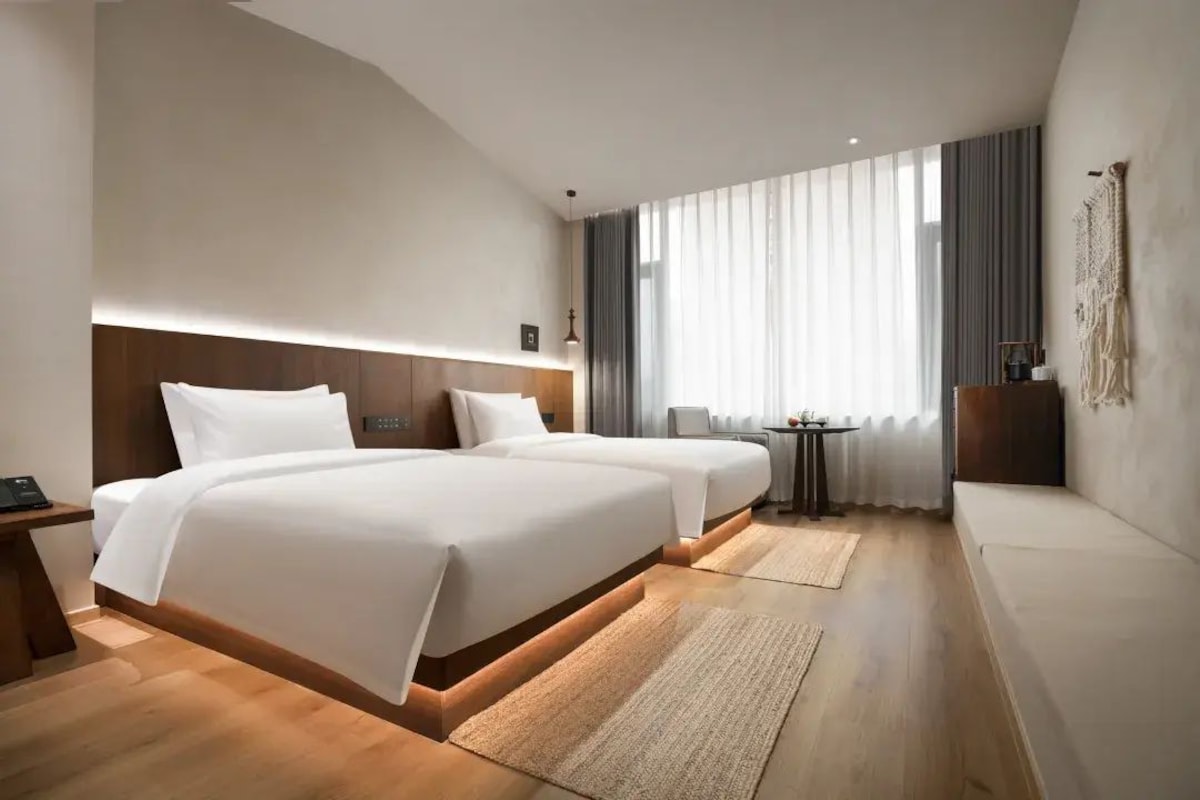 Quarto de hotel com duas camas, uma mesa pequena, um sofá e uma janela grande com cortinas.