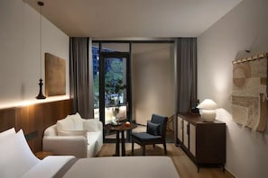 Quarto de hotel com cama, sofá, escrivaninha e cadeira. Há uma janela com cortinas, uma luminária na escrivaninha e um quadro na parede.