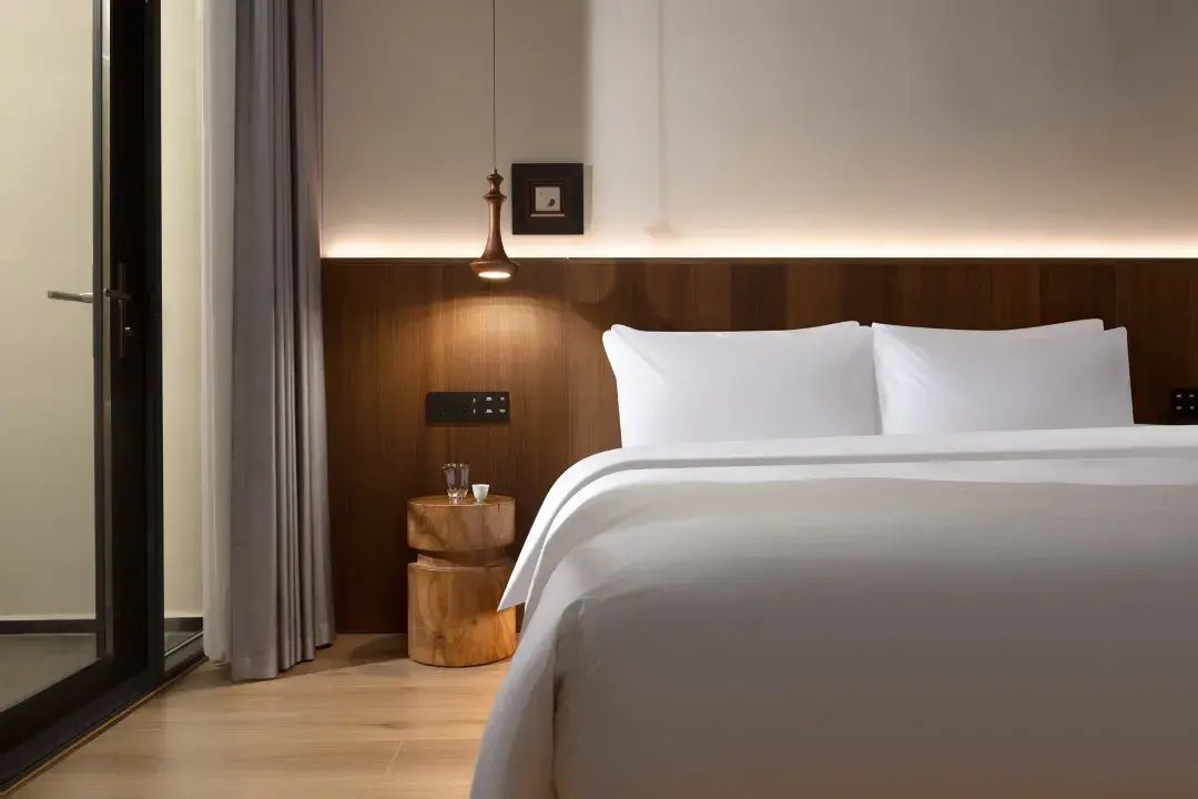Quarto de hotel com cama, mesa de cabeceira com abajur, painel de cabeceira de madeira e quadro pendurado na parede.