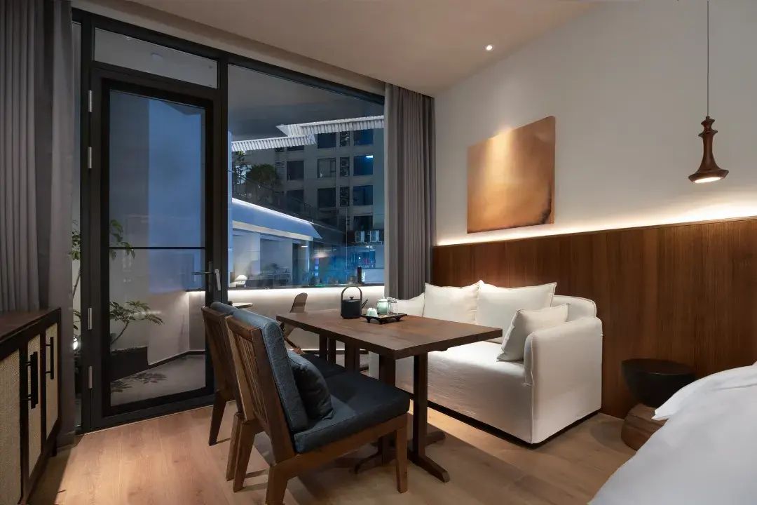 A modern hotel room with a dining area, a sofa, a bed, and a large window offering a view of the cityscape.