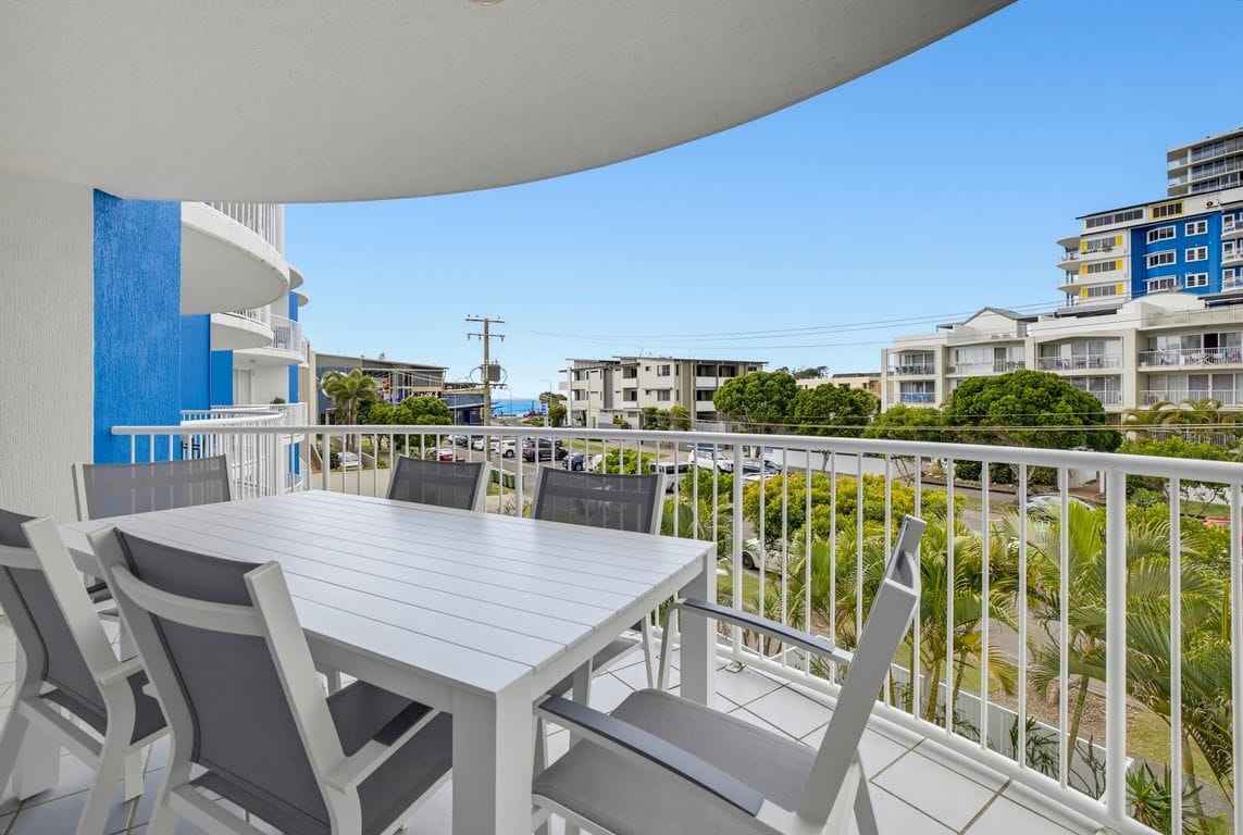 A balcony with a white table and chairs, overlooking a residential area with multi-story buildings and greenery.