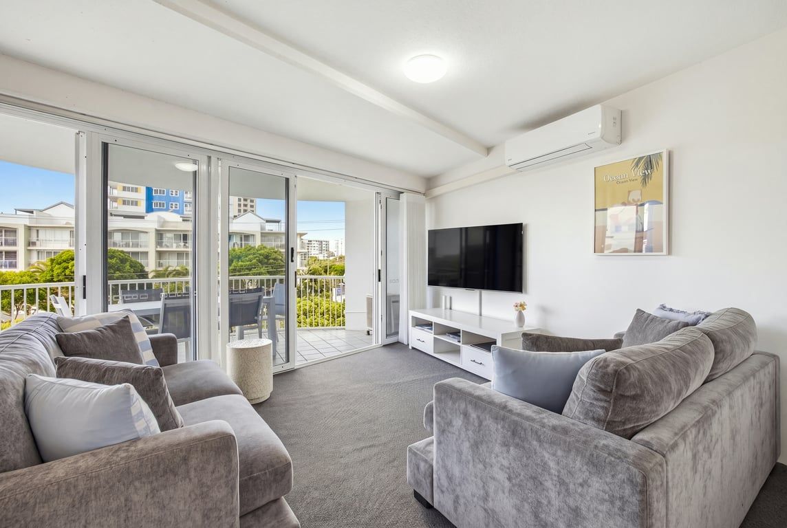 En moderne stue med en grå sofasæt, et fladskærms-tv og en balkon med udsigt til bygninger.