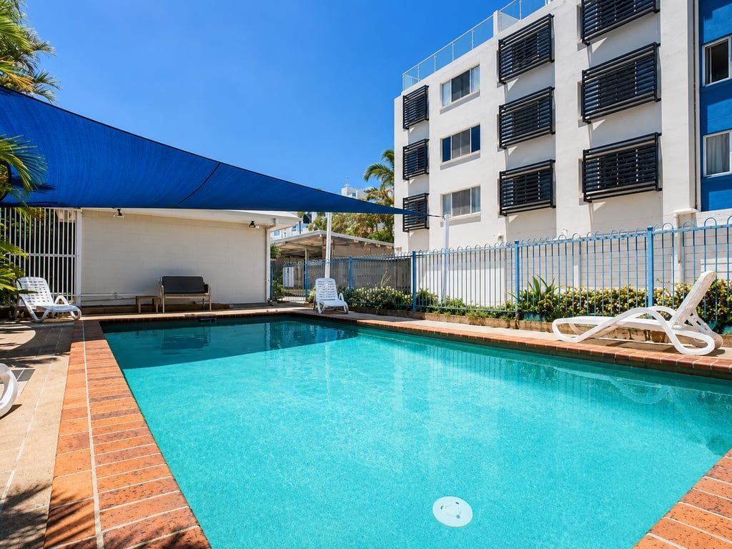 Uma piscina com espreguiçadeiras, um toldo azul e um prédio com varandas.