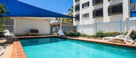 A swimming pool with lounge chairs, a blue canopy, and a building with balconies.