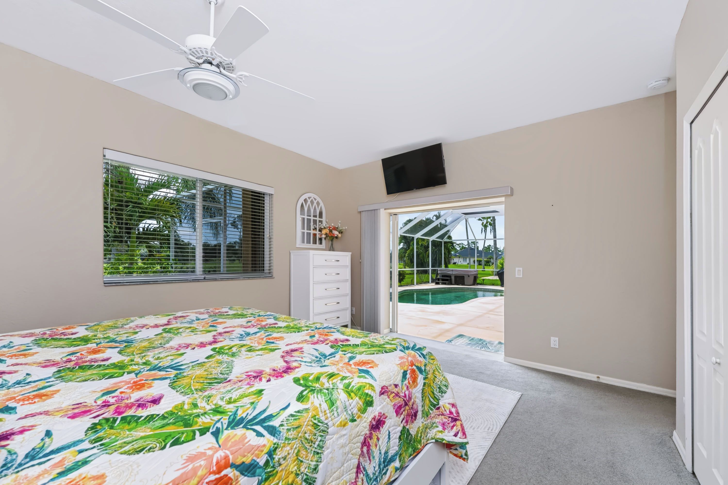 A bedroom with a bed, a ceiling fan, a TV, a window with blinds, and a view of a swimming pool.