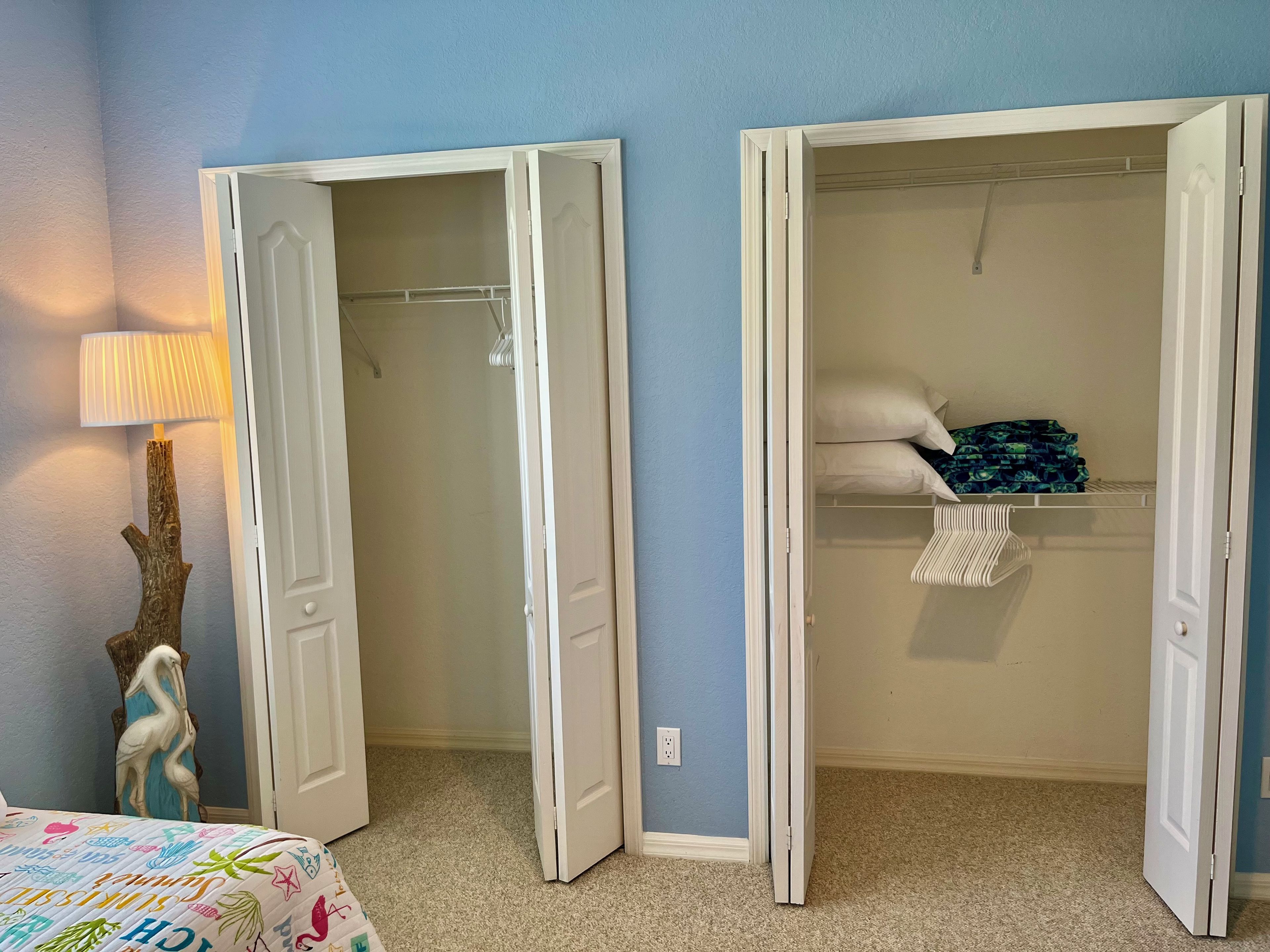 Een slaapkamer met een bed, nachtkastje, lamp en een inbouwkast met hangende kleding.