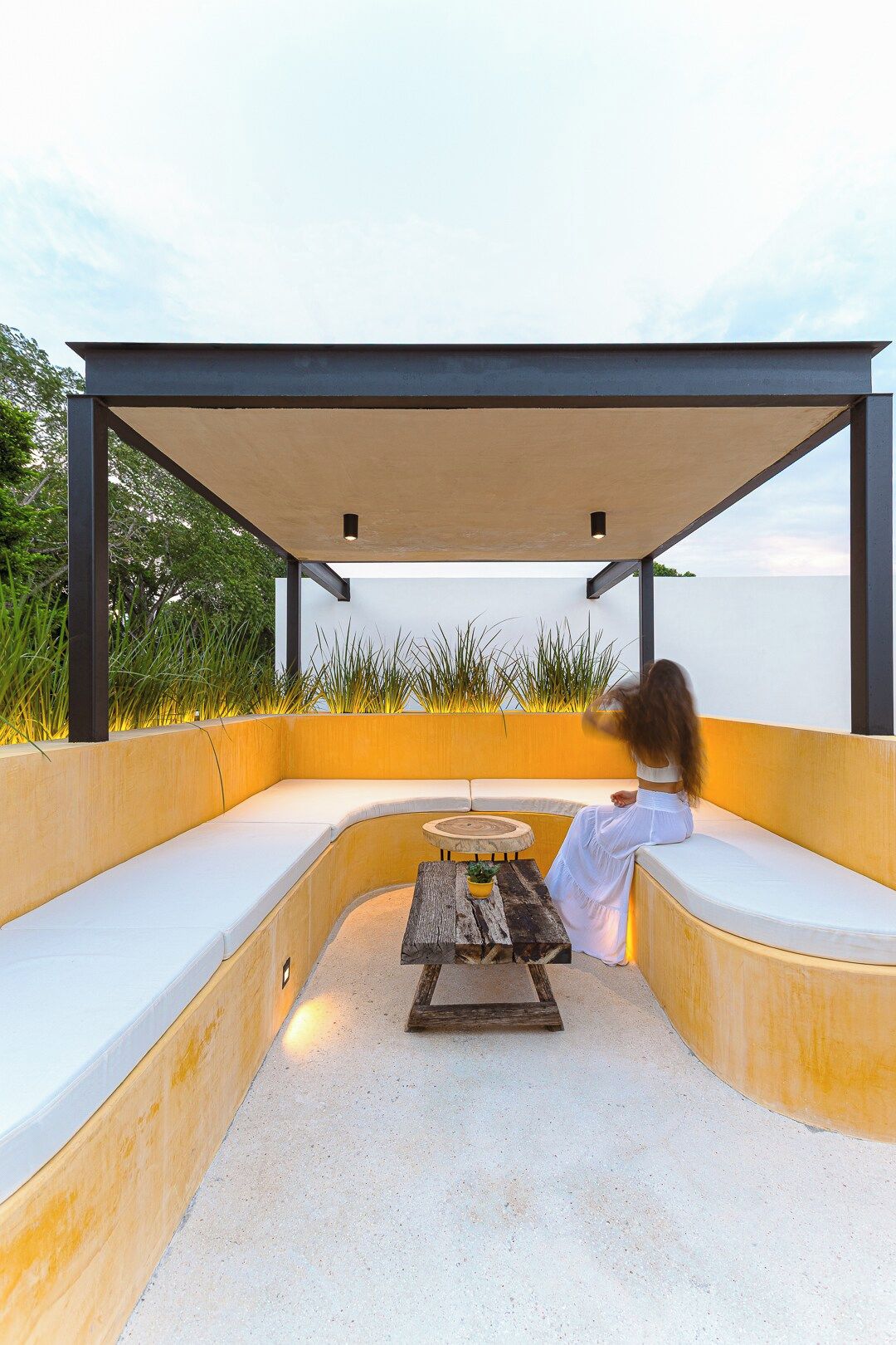 Un espace extérieur aménagé avec une table et des bancs, entouré d’un petit muret et surmonté d’une pergola.