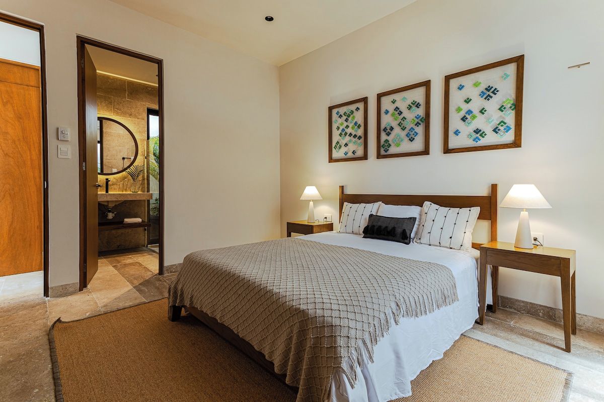 Une chambre d’hôtel avec un grand lit, deux tables de chevet avec des lampes, une tête de lit en bois et trois œuvres d’art encadrées accrochées au mur.