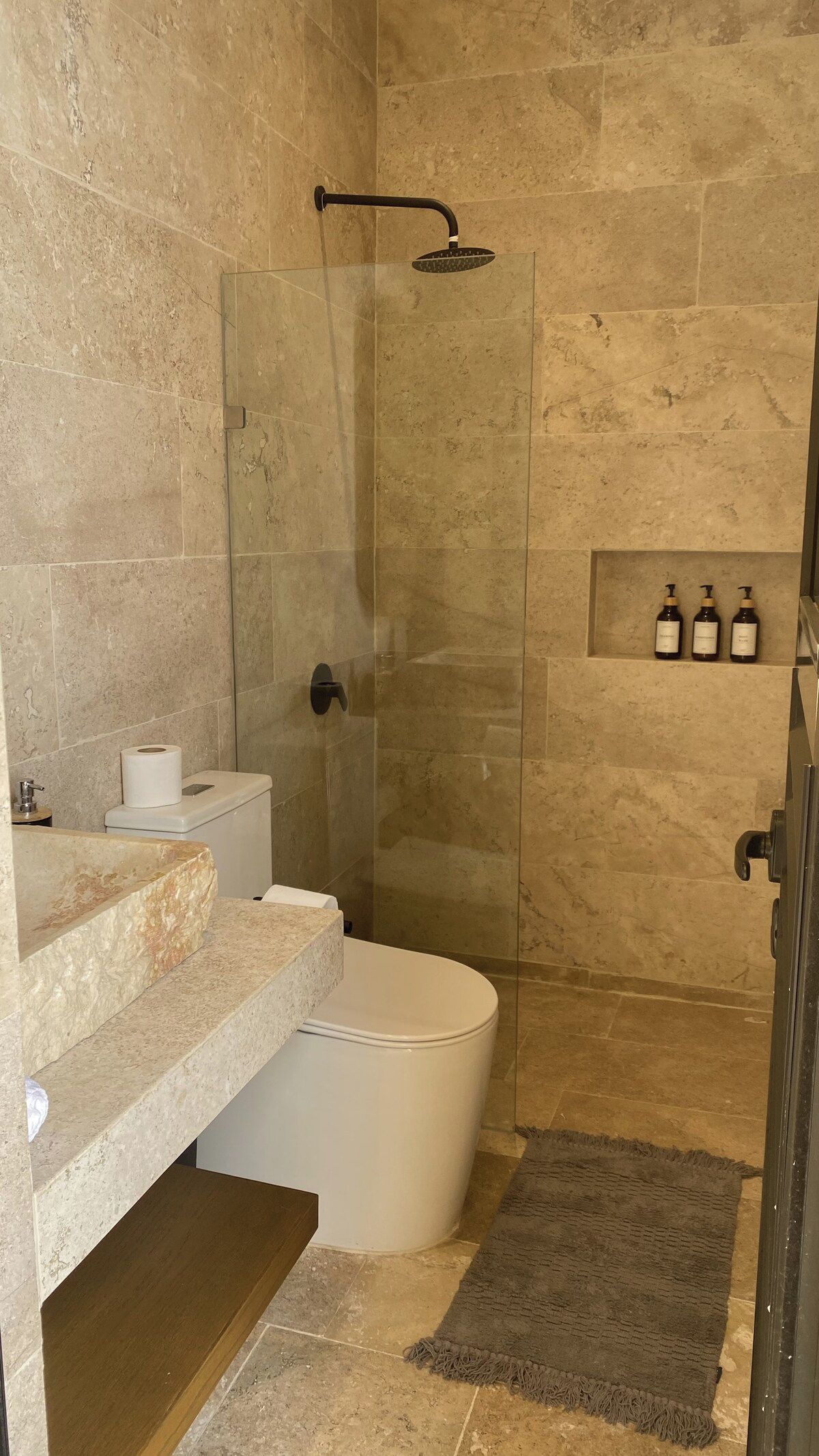 Ein Badezimmer mit einer gläsernen Duschkabine, einer Toilette, einem Waschtisch mit Marmorplatte und einer kleinen Ablage für Pflegeprodukte.