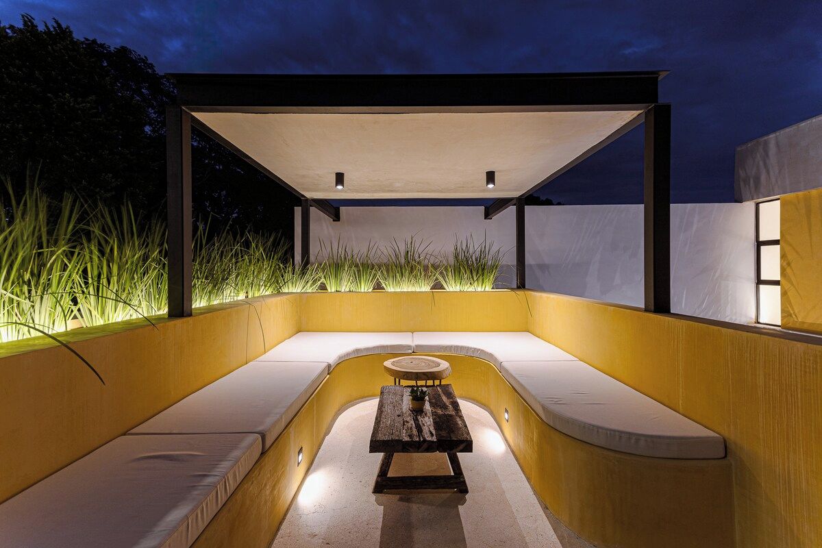 Un espace extérieur aménagé avec une table et des bancs, entouré d’un mur jaune et d’une partie abritée.