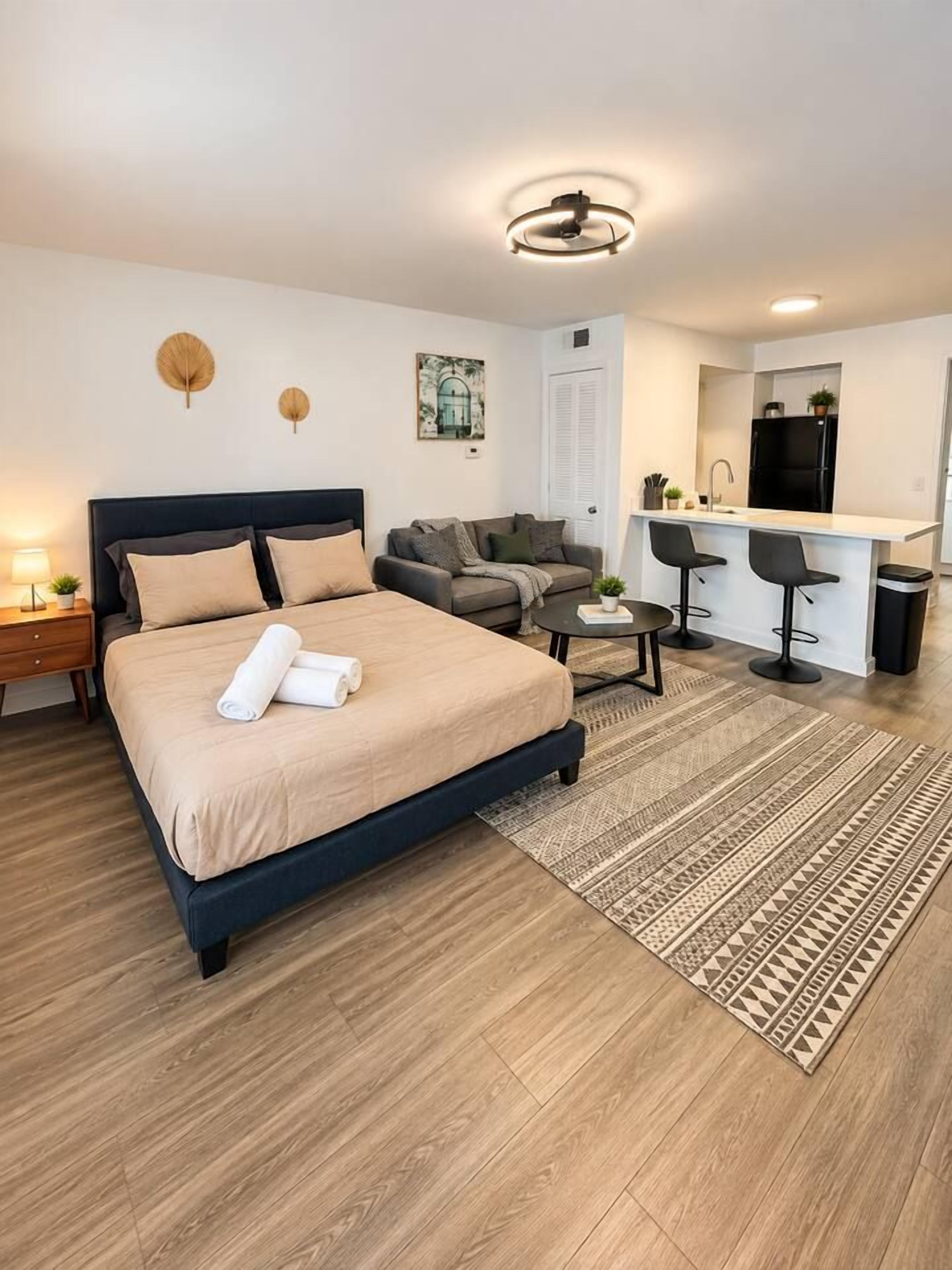 Une chambre d’hôtel moderne dotée d’un grand lit, d’un coin salon avec un canapé et des fauteuils, d’un petit coin cuisine et d’un luminaire au plafond.