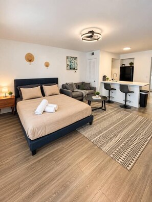 A modern hotel room with a large bed, a sitting area with a sofa and chairs, a small kitchen area, and a ceiling light fixture.