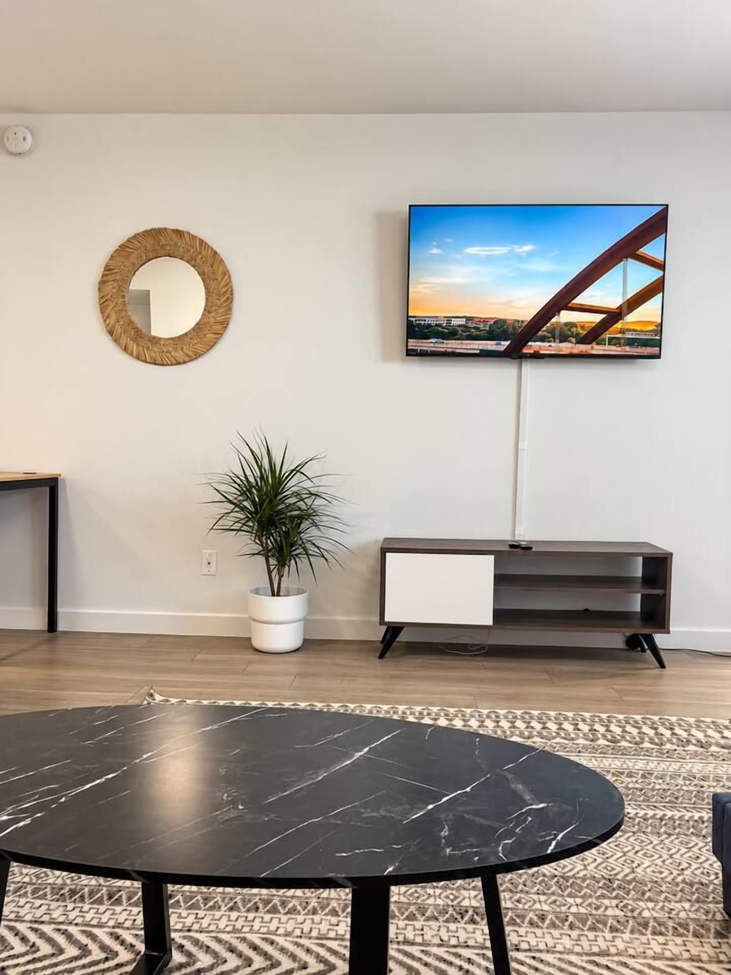 Un salon avec une table ronde noire, une plante en pot blanche, un meuble TV en bois et un tableau encadré accroché au mur.