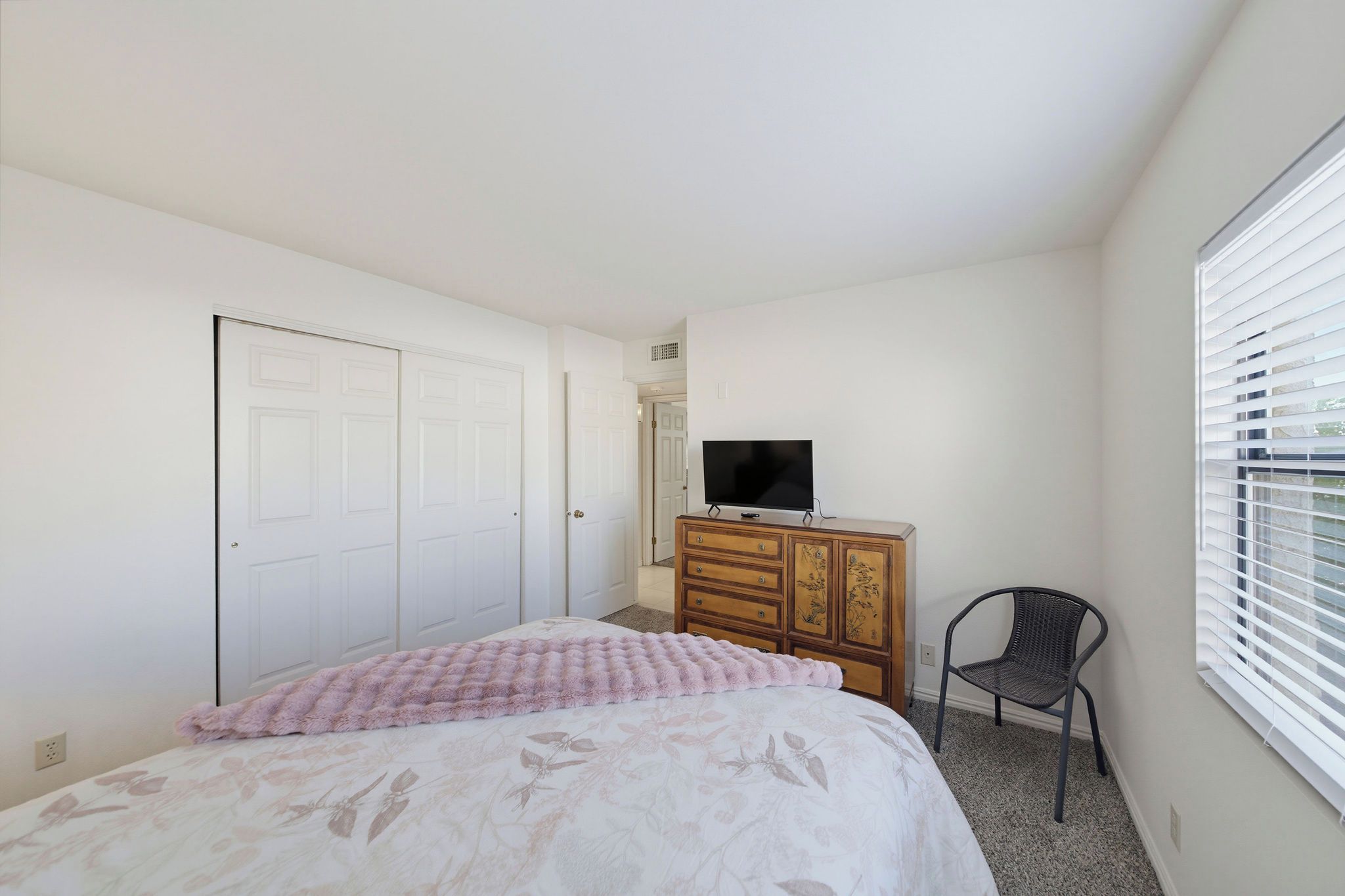 A bedroom with a bed, a dresser, a television, a chair, and a window with blinds.