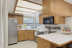 A modern kitchen with wooden cabinets, a stainless steel refrigerator, a microwave, a stove, and a large window providing natural light.