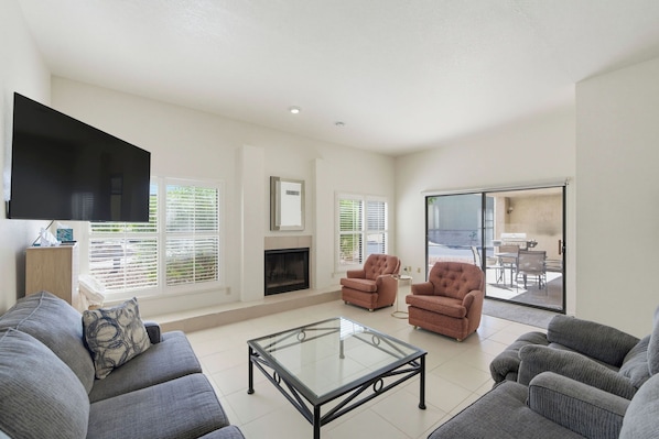 A living room with a flat-screen TV, two armchairs, a sofa, a glass coffee table, and a fireplace.