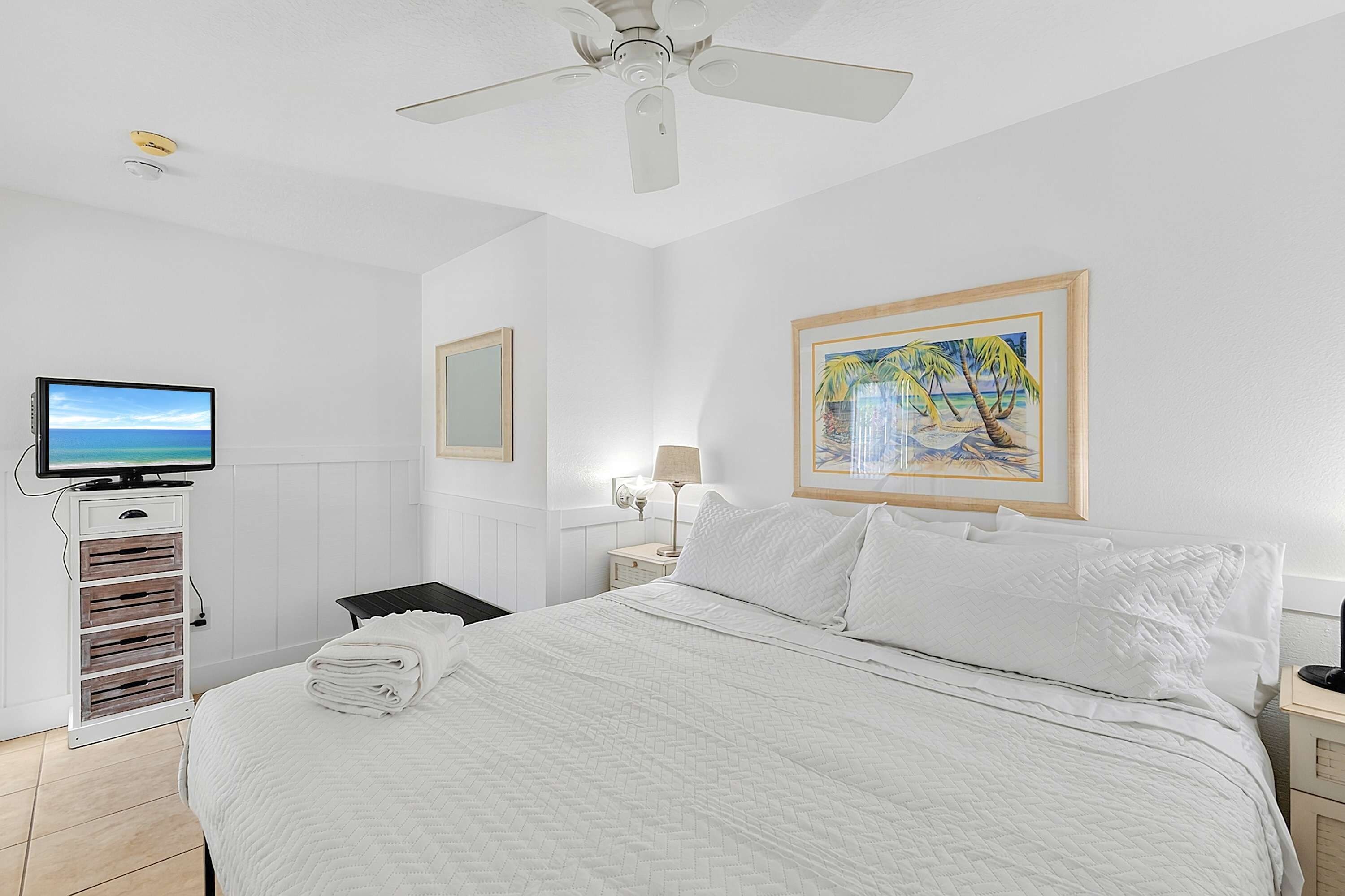 A hotel room with a bed, a television, a painting, and a ceiling fan.