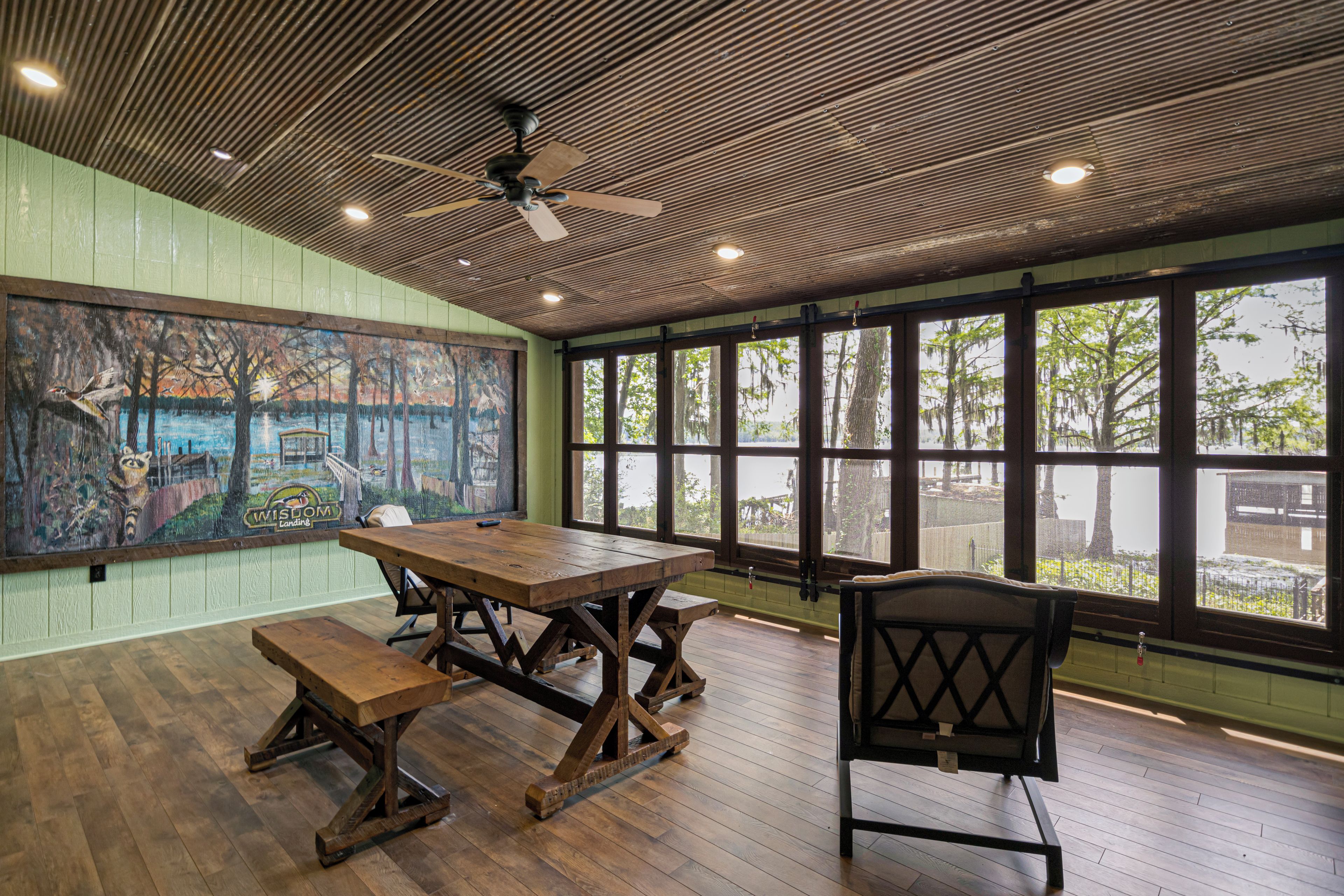 Un espace repas avec une table et des bancs en bois, un ventilateur de plafond et de grandes fenêtres donnant sur la forêt.