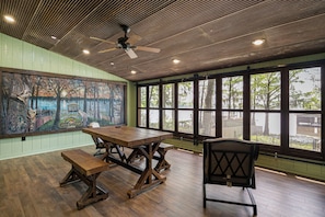A dining area with a wooden table and benches, a ceiling fan, and large windows with a view of trees.