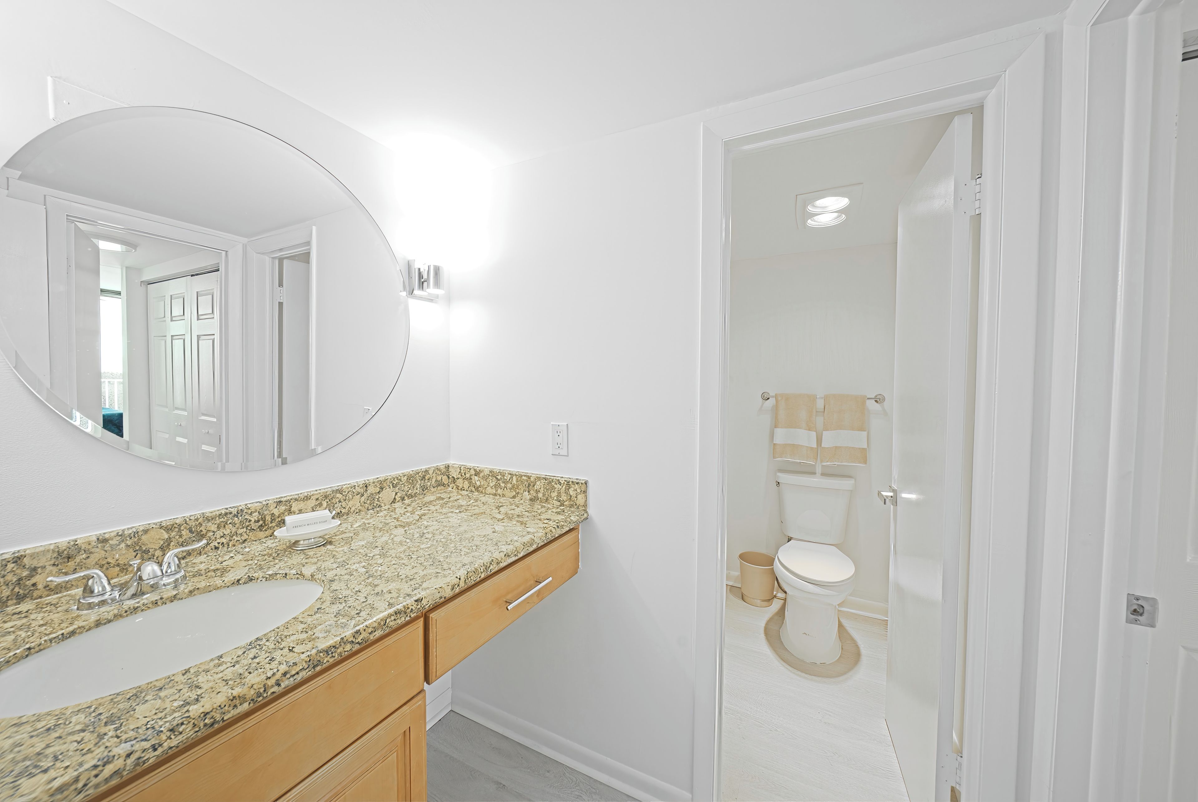 Une salle de bain avec un comptoir en granit, un lavabo, un miroir et des toilettes visibles à travers une porte en verre.