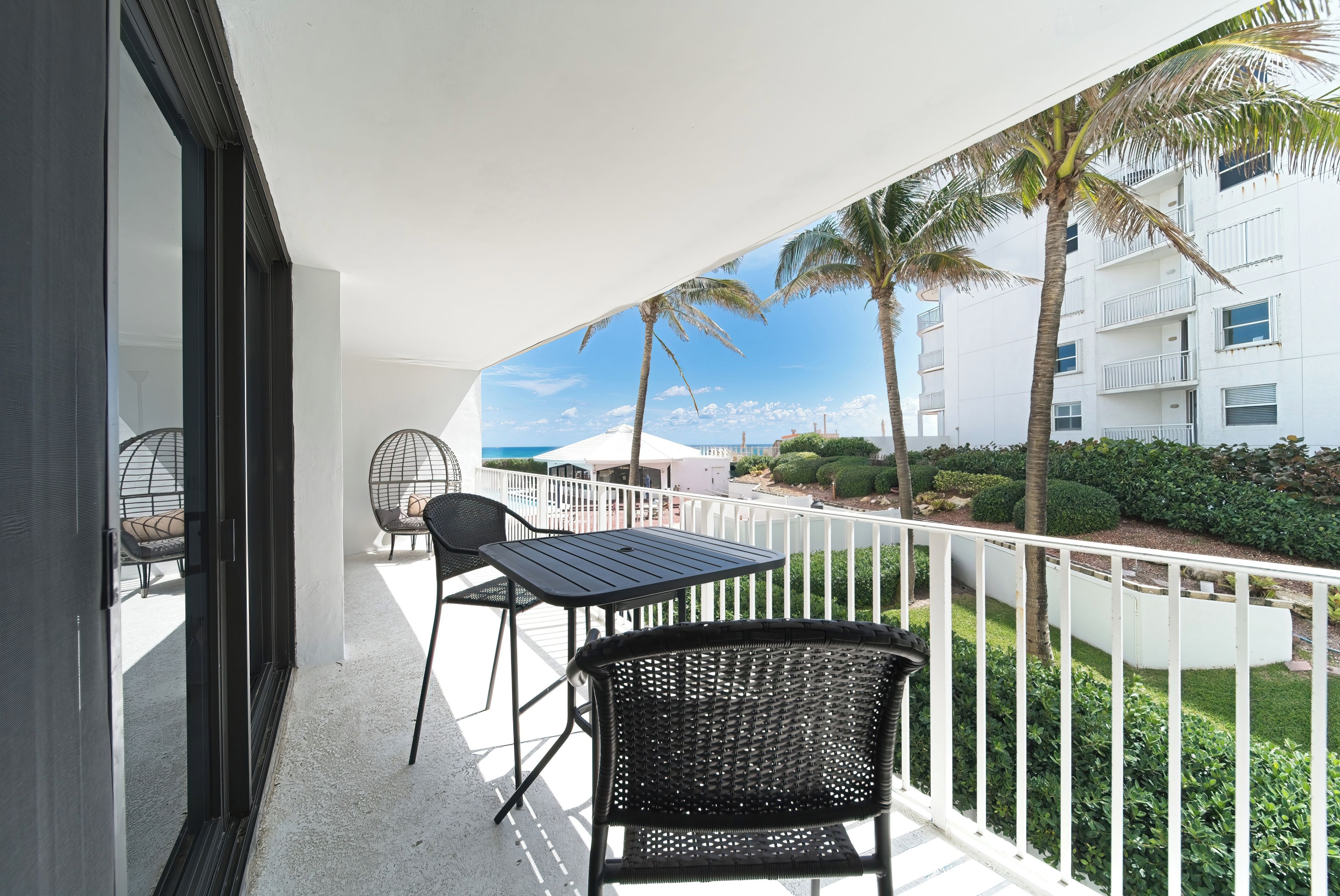 Un balcone con tavolo e sedie, vista su una piscina e palme, con un edificio sullo sfondo.