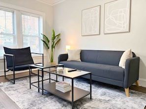 Een moderne woonkamer met een donkerblauwe bank, een zwarte fauteuil, een glazen salontafel en abstracte wanddecoratie.