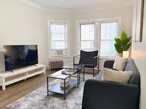 En moderne stue med et fladskærms-tv, et glas sofabord, en sort lænestol, en grå sofa og en stor grøn plante.