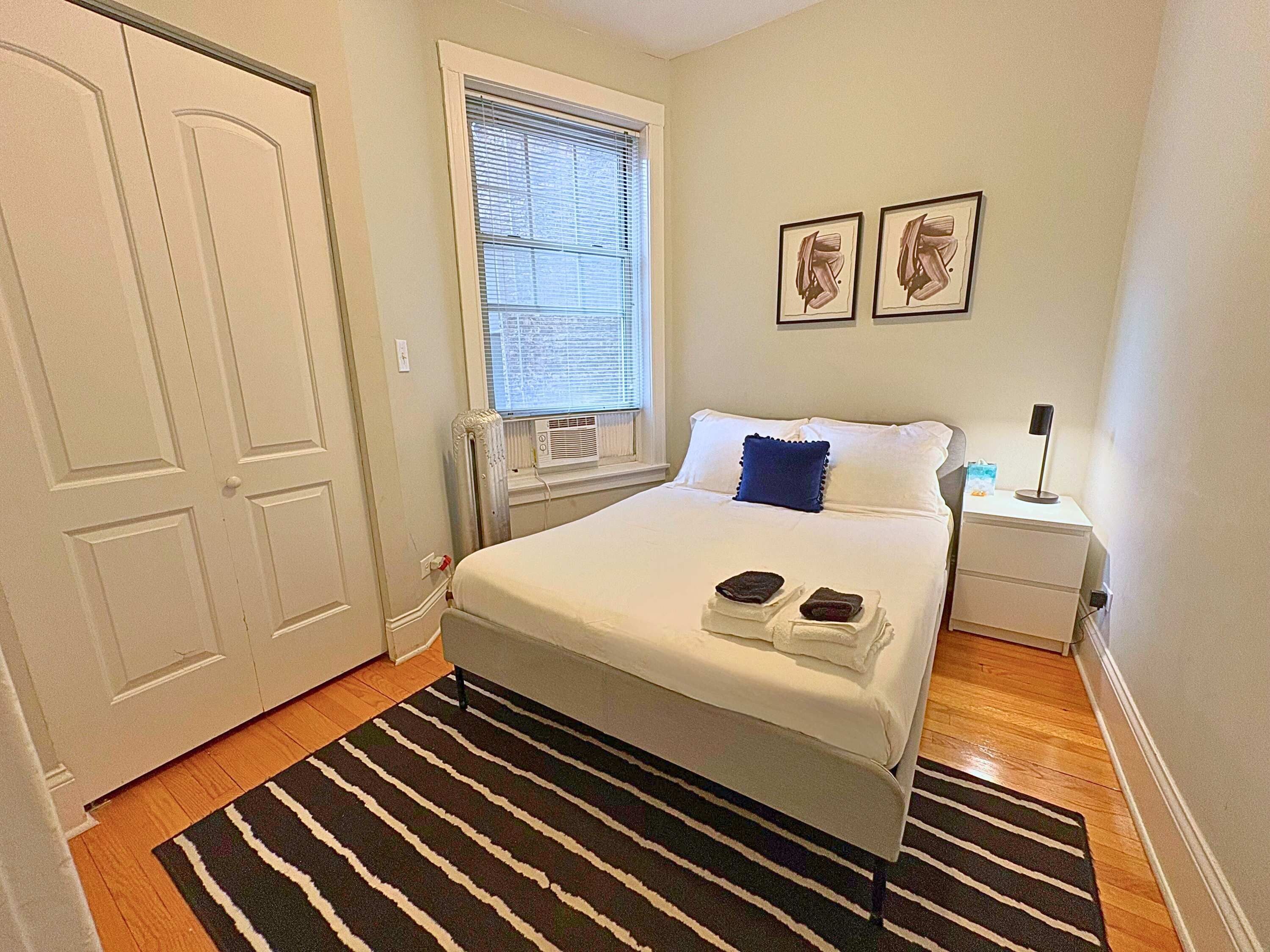 A neatly arranged bedroom with a bed, a bedside table with a lamp, and a window with blinds.