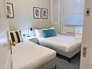 Een hotelkamer met twee bedden, een nachtkastje met een lamp, een raam met jaloezieën en kunst aan de muur.