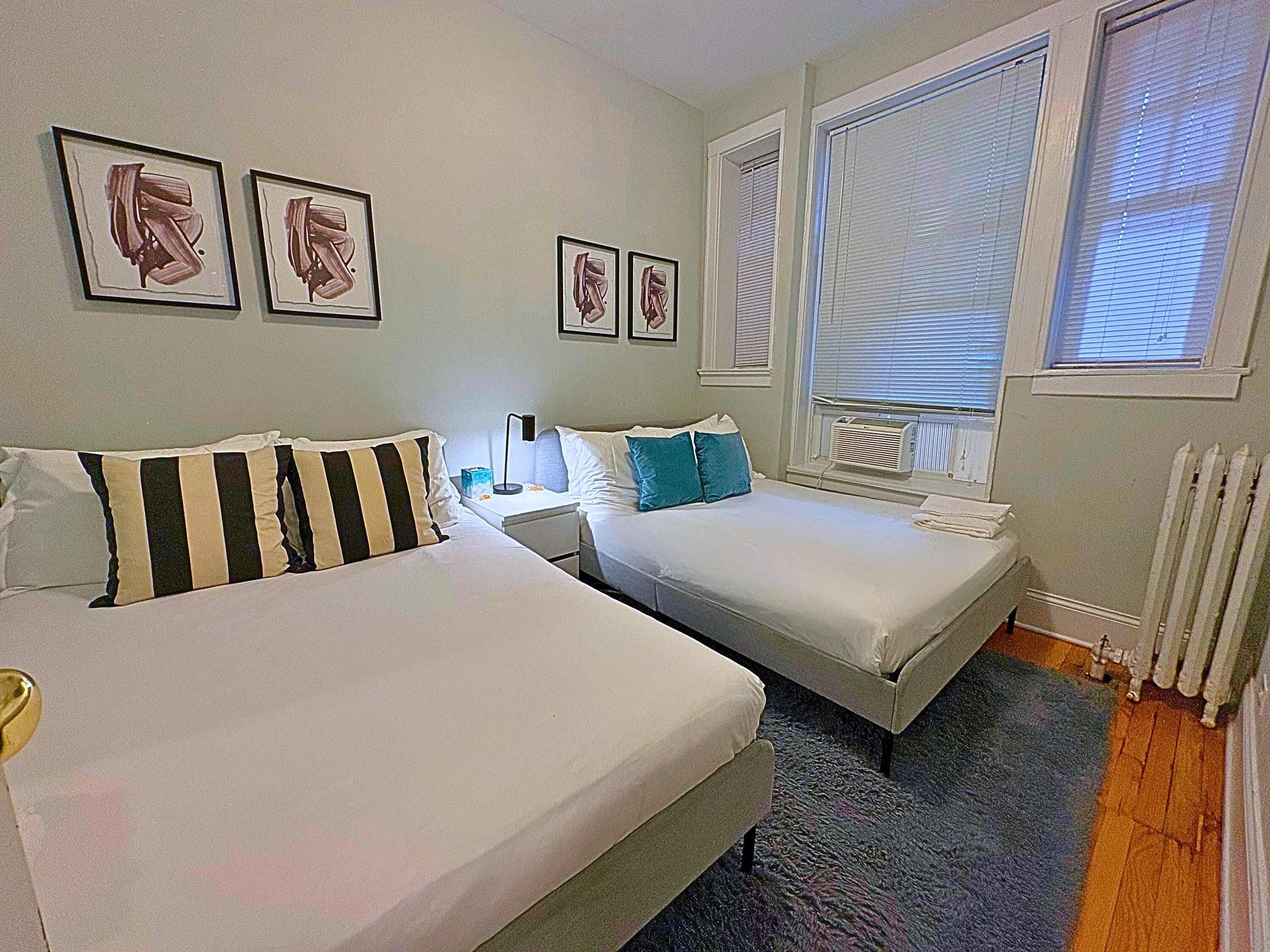 Une chambre d’hôtel avec deux lits, une petite table, une table de chevet et des œuvres d’art encadrées accrochées au mur.