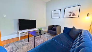 Ein Wohnzimmer mit einem großen Flachbildfernseher, einem blauen Sofa, einem kleinen Tisch mit einer Pflanze und zwei gerahmten Bildern an der Wand.